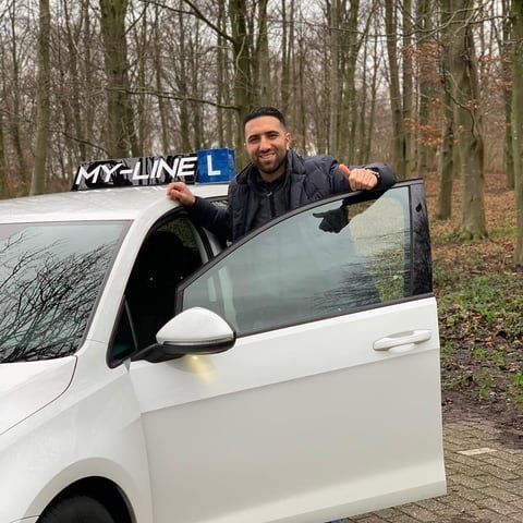 De instructeur van Rijschool MY-Line leunt lachend uit de lesauto met het dakbord