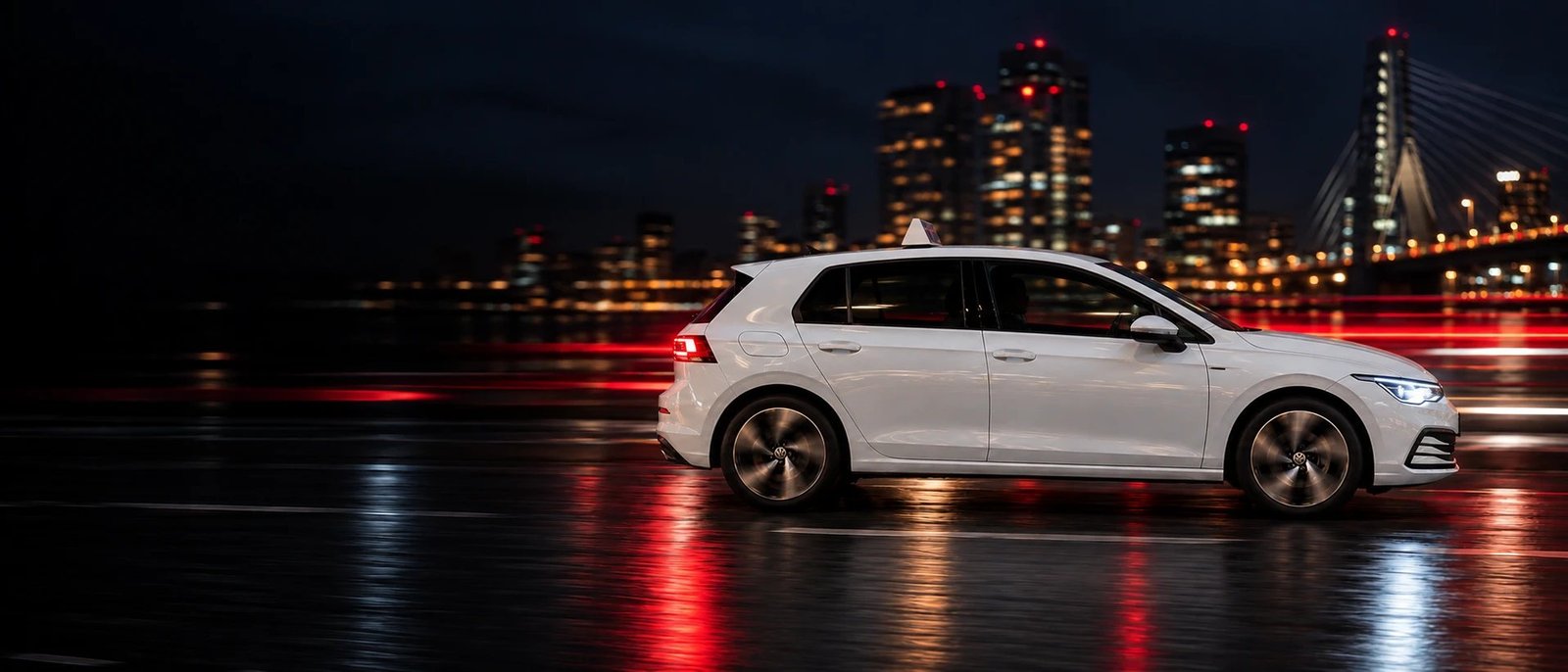 De witte Volkswagen Golf van Rijschool MY-Line met het dakbord rijdt 's avonds over een natte weg, met lichtstrepen erachter