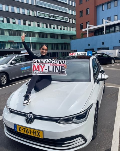 Geslaagde leerling zit met het bord Geslaagd bij MY-Line op de motorkap van de witte lesauto