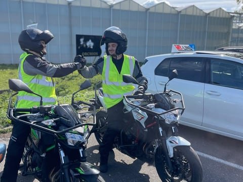 Twee motorleerlingen op de lesmotoren geven elkaar een boks, met de lesauto van MY-Line erachter